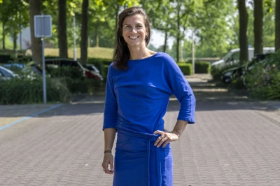 Vrouw in een blauwe jurk staat op een parkeerplaats met bomen op de achtergrond.