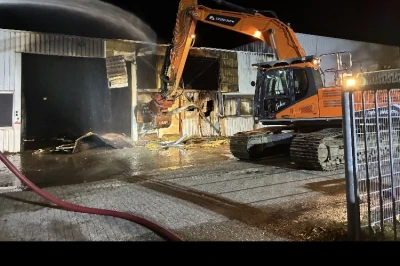 Een graafmachine werkt 's nachts aan een brandlocatie, omringd door waterstralen.