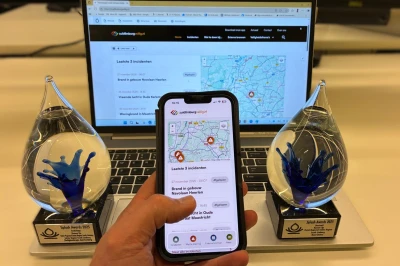 Een hand houdt een smartphone vast voor een laptop met twee trofeeën ernaast.