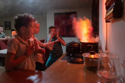 Kinderen kijken geschrokken naar rook en vlammen uit een kookpot in de keuken.