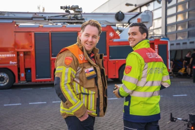 Twee brandweermannen staan glimlachend bij een brandweerauto.