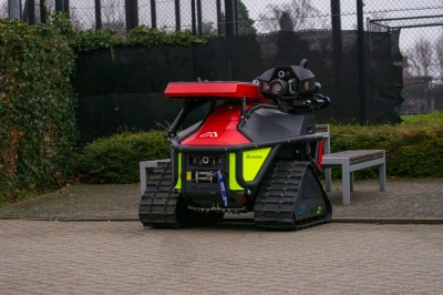 De blusrobot is een rupsvoertuig met een rode kap en blusarm, geparkeerd op een bestrate ondergrond nabij een hek.