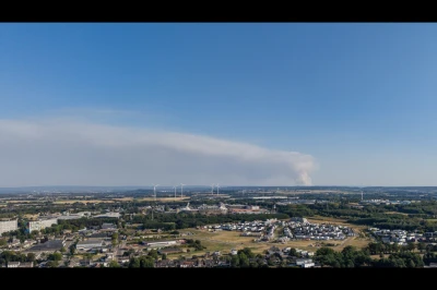 Luchtfoto van een uitgestrekte stad met rookpluim in de verte en een heldere blauwe lucht.