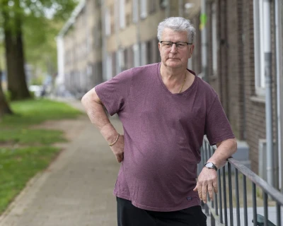 Man met grijs haar in een paarse shirt leunt tegen een hek in een stedelijke omgeving.