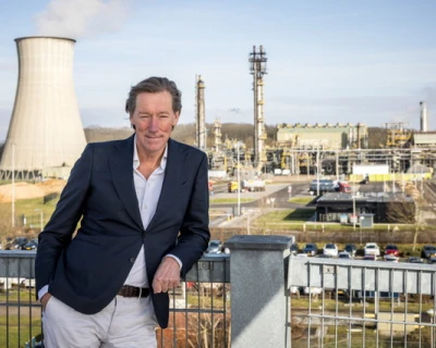 Een man in een donkerblazer staat op een balustrade, met een industriegebied op de achtergrond.