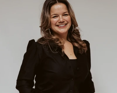 Vrouw met een glimlach, gekleed in zwart, staat met handen in haar zakken tegen een neutrale achtergrond.
